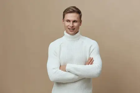 Молодой улыбающийся светлокожий мужчина в белом вязаном свитере с высоким воротом стоит, скрестив руки, глядя в сторону, на бежевом фоне