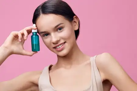Улыбающаяся азиатская модель демонстрирует зеленую сыворотку на розовом фоне, подчеркивая красоту, уход за кожей и оздоровительные продукты для сияющего цвета лица