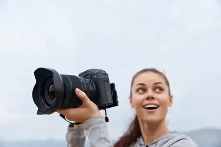 Счастливая молодая женщина высоко поднимает свой фотоаппарат, запечатлевая момент вдохновения на открытом воздухе