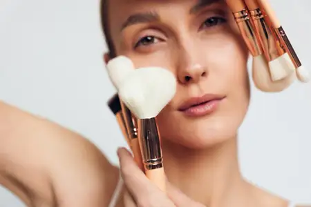 Сцена изображает безмятежную модель во время ретуши макияжа: мягкое освещение подчеркивает гладкую кожу, нейтральные тона в палитре и сосредоточенную, тихую атмосферу
