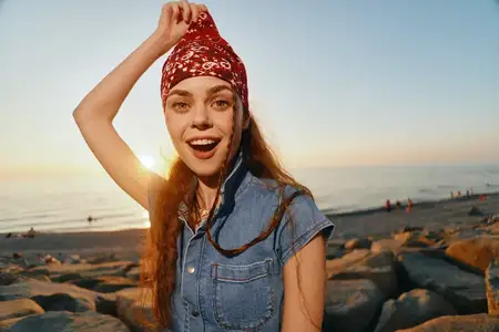 Женщина в стиле современного богемно-вестерн денима ярко улыбается на пляже во время заката, передавая беззаботную и живую атмосферу плёночной цветокоррекции