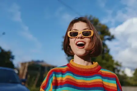 Женщина улыбается в полосатом свитере и желтых солнцезащитных очках, молодая взрослая с коротким бобом, радостная смеющаяся поза, стоя на улице под голубым небом для истории о путешествиях, образе…