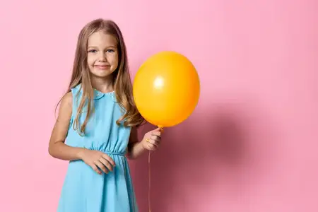 Весёлая маленькая девочка в голубом платье держит жёлтый воздушный шар на ярко-розовом фоне