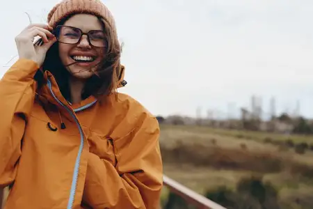 Женщина на открытом воздухе в стиле лайфстайл: улыбается в куртке и вязаной шапке-бини, видны очки, отвечает на звонок по смартфону, осенний пейзаж, мягкое размытие, открытое небо, естественный…