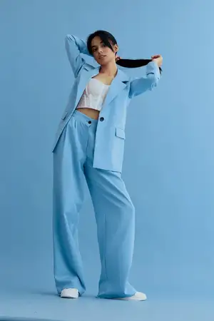 Молодая женщина в синем oversized костюме и белом кружевном укороченном топе уверенно стоит, держась за свой хвост, на фоне бесшовного синего фона, подчеркивающего модную повседневную элегантность