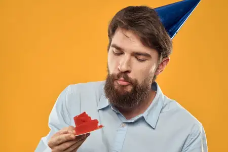 Мужчина с тортом на жёлтом фоне, в праздничном колпаке на день рождения, кадр обрезан