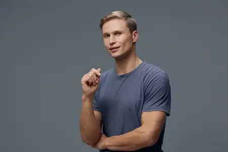 Уверенный молодой улыбающийся мужчина в повседневной синей футболке стоит, скрестив руки, и смотрит в камеру на однотонном сером фоне, с акцентом на концепцию психического здоровья и гаджетов…