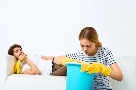 Женщина с ведром в резиновых перчатках убирается дома, а её муж лежит на диване