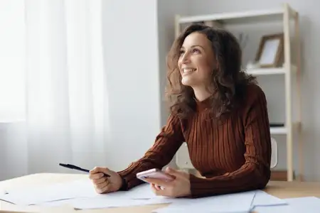 Веселая, смеющаяся, счастливая, кудрявая, милая женщина сидит за столом с бумагами и документами, работая из дома