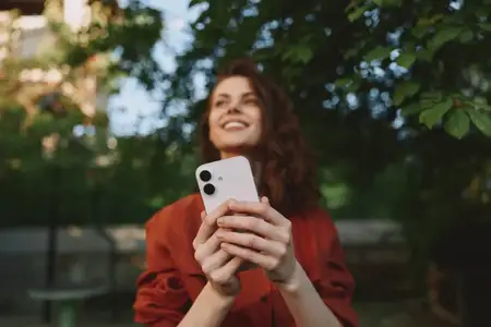 Улыбающаяся женщина на улице запечатлевает искренний момент с телефоном в обрамлении деревьев и мягкого солнечного света