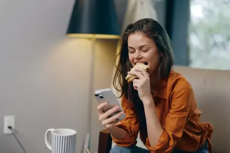 Молодая женщина в повседневной оранжевой рубашке наслаждается вкусным сэндвичем, одновременно глядя в смартфон, в уютной домашней обстановке, что способствует пропаганде здорового питания