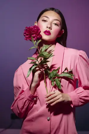Элегантная модель-женщина в ярко-розовом пиджаке позирует с пышным цветком пиона на темно-фиолетовом фоне в красиво стилизованной креативной модной редакции, подчеркивающей красоту и грацию