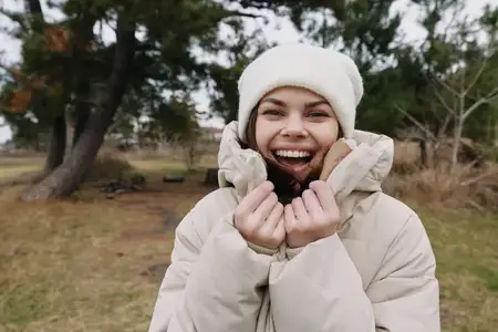 Радостный портрет женщины в зимней одежде на улице: она укутана в пуховик и шапку-бини, наслаждается прохладным днём в парке на фоне природного пейзажа