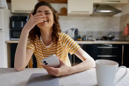 Жизнерадостная женщина дома на кухне, смартфон в руке, расслабленное настроение, откровенная сцена в помещении, демонстрирующая повседневный образ жизни и домашнюю атмосферу со светом и теплом