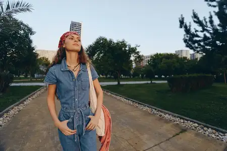 Стильная женщина, ведущая активный образ жизни, в современном богемно-вестерн дениме уверенно стоит на дорожке парка в окружении зеленых деревьев и городских зданий