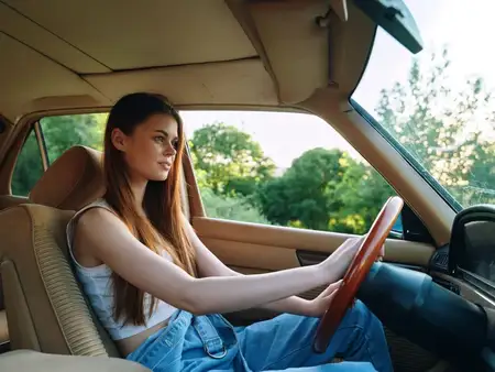 Молодая женщина с длинными волосами ведет классический автомобиль, излучая уверенность и чувство свободы на фоне яркой зеленой природы