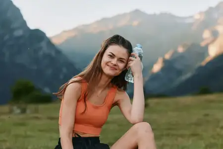 Жизнерадостная женщина наслаждается моментом на природе на фоне величественных гор, в спортивной одежде, потягивая воду, излучая энергию, здоровье и свободу