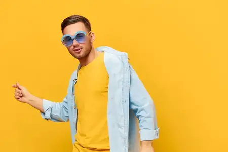 Весёлый загорелый красивый мужчина в синей рубашке и модных солнцезащитных очках танцует и поёт, позируя изолированно на оранжево-жёлтом студийном фоне