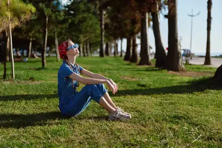 Женщина в стиле современный бохо-вестерн, одетая в джинсовую уличную одежду, расслабленно сидит на траве под деревьями, наслаждаясь солнечным днём в стильных солнцезащитных очках и красной бандане…