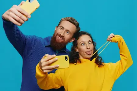 Забавная пара — женщина и мужчина с телефонами в руках делают селфи с кривой улыбкой, веселятся на синем фоне