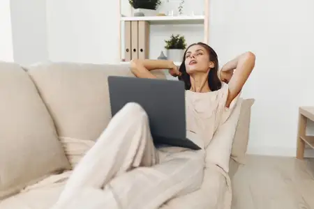 Улыбающаяся женщина работает за ноутбуком в уютной гостиной в окружении современных технологий и устройств для досуга