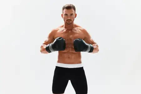 Спортивный мужчина, бодибилдер, позирует в боксерских перчатках с обнаженным торсом и рельефным прессом в полный рост на фоне, занимаясь боксом и единоборствами