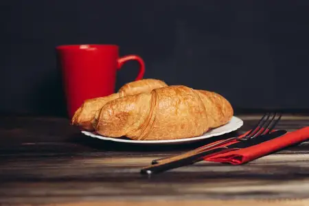 Свежий круассан на тарелке, кухонная утварь, чашка кофе, завтрак