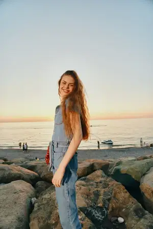 Счастливая женщина в современном джинсовом уличном наряде в стиле бохо-вестерн стоит на камнях у океана в теплых тонах кинопленки на закате, вызывая ощущение расслабленной летней атмосферы