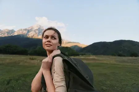 Молодая женщина с рюкзаком стоит на широком лугу с холмистыми холмами и горами на заднем плане, одетая в легкий нейтральный топ, наслаждаясь природой и уединением
