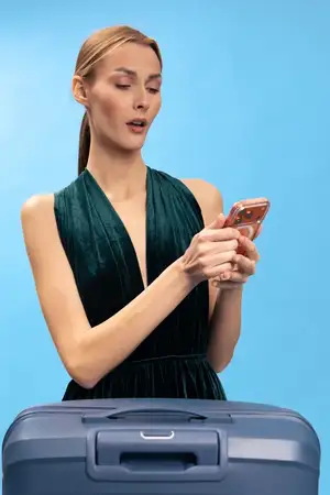 Элегантная женщина с хвостиком, держащая смартфон и выглядящая удивленной, рядом с синим чемоданом на светло-голубом фоне