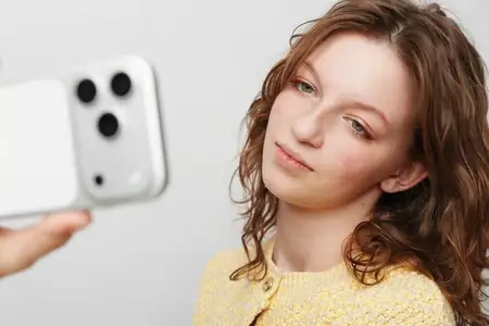 Женщина с вьющимися волосами в желтом свитере держит смартфон для селфи