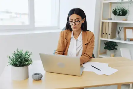 Деловая женщина в бежевом костюме и белой рубашке в очках работает в офисе за столом с ноутбуком, занимаясь документами и бухгалтерией