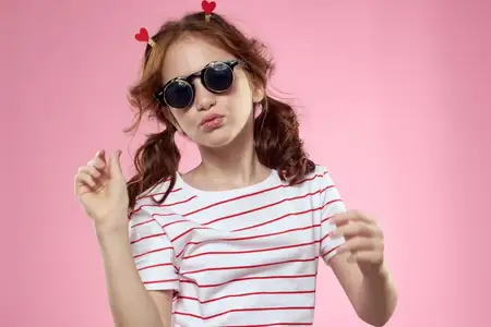 Маленькая девочка в модных очках и с зажимами для волос, её эмоции на розовом фоне с сердечками