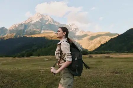Молодая женщина в бежевой рубашке и шортах несет черный рюкзак во время похода по бескрайнему горному лугу под ясным небом, наслаждаясь природой и приключениями