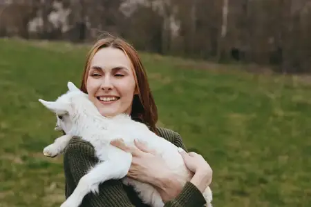 Яркий портрет женщины с козлёнком на открытом воздухе, откровенный момент на зелёном лугу, тёплый свет, игривая привязанность, деревенская атмосфера, простая здоровая жизненная сцена