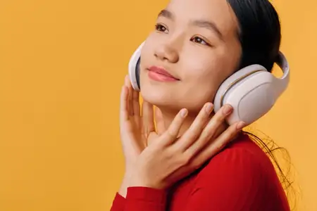 Улыбающаяся женщина в больших белых наушниках слушает музыку с умиротворенным выражением лица, касаясь гарнитуры рукой, на ярком желтом фоне в красной блузке с длинным рукавом