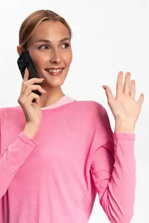 Улыбающаяся женщина в розовом свитере пользуется смартфоном и машет рукой в знак приветствия