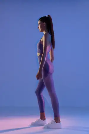 Атлетичная спортивная фитнес-женщина в фиолетовом спортивном костюме стоит в профиль в мягко освещенной чистой студийной обстановке, подчеркивая преданность делу и здоровый образ жизни