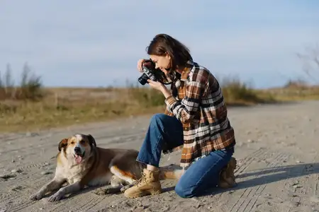 Уличная сцена: фотограф с камерой фокусируется на спокойной собаке, отдыхающей на земле, ветер в волосах, повседневная одежда, деревенская обстановка, природный фон и искренний творческий момент