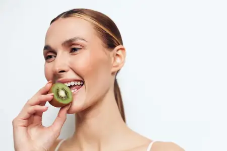 Счастливая женщина с безупречной кожей откусывает кусочек свежего киви на однотонном белом фоне