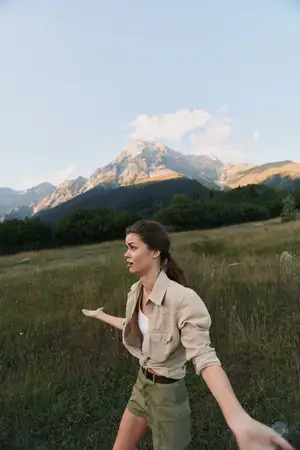 Молодая женщина с длинными каштановыми волосами в бежевой куртке и зеленых шортах стоит на травянистом поле на фоне гор под голубым небом