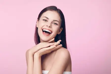 Портрет счастливой молодой женщины, которая улыбается, приложив руки к лицу, на розовом фоне