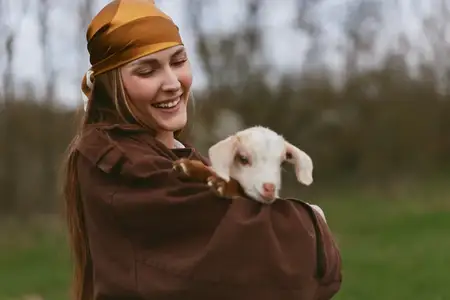 Молодая женщина в коричневой куртке и оранжевой косынке нежно обнимает козленка на улице в окружении мягкой зелени, выражая радость и связь в искренней лайфстайл-сцене