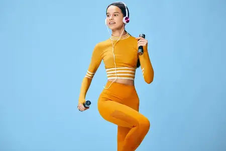 Спортивная спортсменка в ярком оранжевом костюме демонстрирует упражнения на мобильность и кардио, с беспроводными наушниками, на студийном фоне, подчеркивая силу, здоровье и активный образ жизни