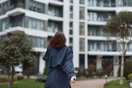 Она стоит спиной, прогуливаясь по мощёной дорожке, обрамлённой деревьями и стеклянными зданиями, что вызывает ощущение спокойного городского образа жизни при мягком дневном свете
