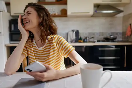 Непринужденная домашняя сцена: улыбающаяся женщина в полосатой футболке сидит за кухонным столом и пользуется телефоном