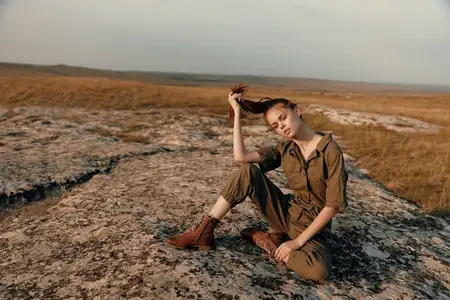 Молодая женщина сидит на камне в безмятежном поле, положив руки на голову, и размышляет, созерцая природу и красоту