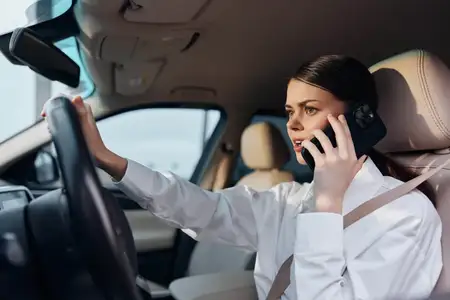 Женщина сидит на водительском сиденье автомобиля, одной рукой держась за руль, а другой разговаривая по мобильному телефону