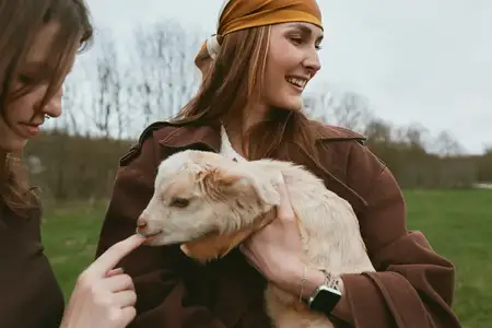 Молодая женщина в коричневой куртке и платке держит белого козлёнка, а другой человек нежно касается носа животного в естественной природной обстановке весной