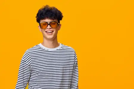 Веселый улыбающийся мужчина с кудрявыми волосами и в солнечных очках, одетый в полосатую рубашку, стоит на ярком оранжевом фоне, излучая юношескую энергию и концепцию повседневной летней моды